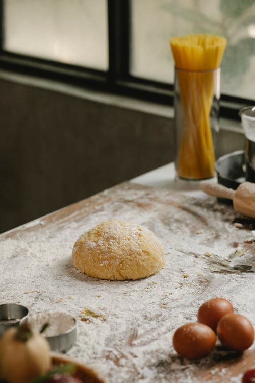 From above ball of dough placed on table covered with flour near raw spaghetti and eggs in kitchen while cooking process