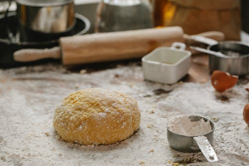 Ball of raw dough placed on table sprinkled with flour near rolling pin dishware and measuring cup in kitchen on blurred background