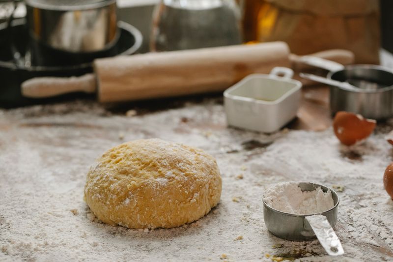 Ball of raw dough placed on table sprinkled with flour near rolling pin dishware and measuring cup in kitchen on blurred background