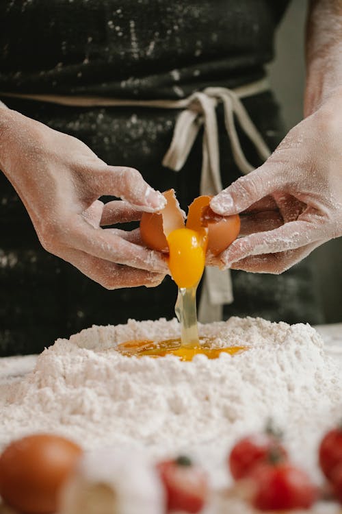 Unrecognizable person wearing messy apron breaking egg into heap of flour while preparing dough at table with tomatoes  in kitchen