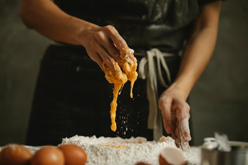Unrecognizable woman in apron kneading dough with eggs and flour on table in kitchen