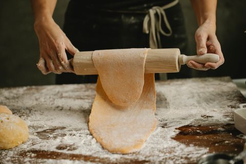 Hands lifting a thin sheet of pasta dough over a wooden rolling pin on floured table