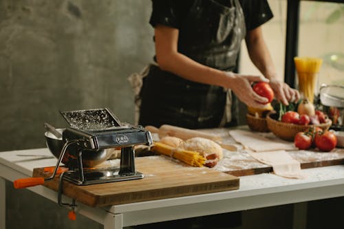 Crop unrecognizable housewife in apron standing near table with dough and vegetables while cooking in kitchen
