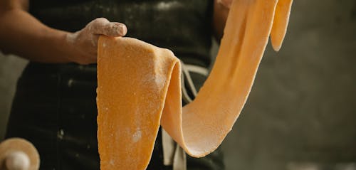 Crop anonymous cook demonstrating soft thin sheet of pasta dough with flour on gray background