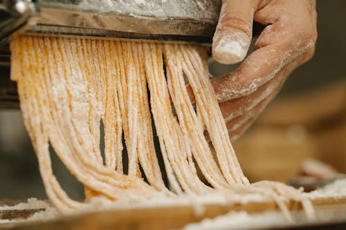Crop unrecognizable chef preparing spaghetti from uncooked dough with flour using pasta rolling machine in kitchen
