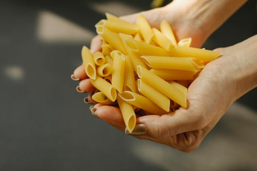 Unrecognizable female demonstrating heap of uncooked penne rigate pasta with ridged surface in hands while standing in kitchen with sunlight