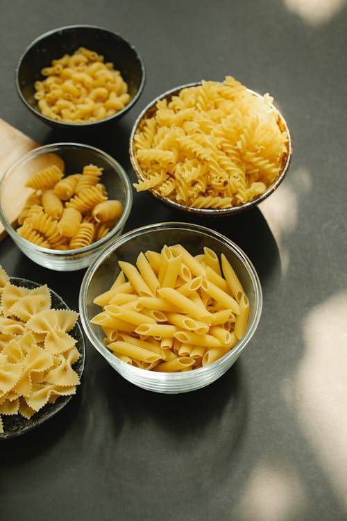 Free From above bowls of penne rigate and fusilli pasta placed on dark table with uncooked farfalle radiatori and elbows types of macaroni Stock Photo