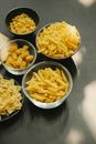 From above bowls of penne rigate and fusilli pasta placed on dark table with uncooked farfalle radiatori and elbows types of macaroni