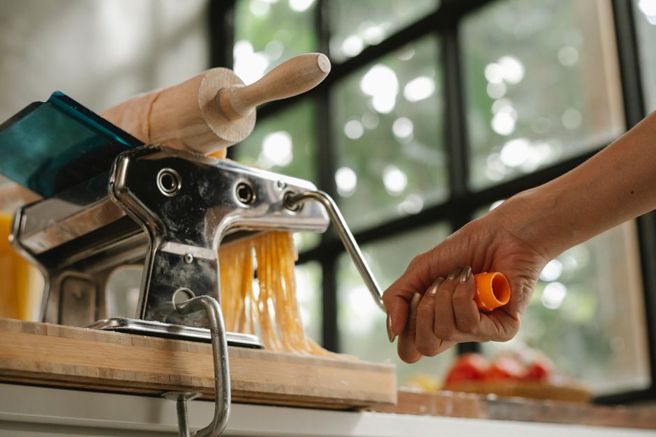 From below of anonymous female cook turning handle of pasta maker machine to make pasta in kitchen on blurred background