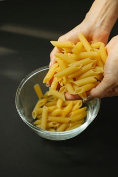 Unrecognizable female cook pouring yellow uncooked penne rigate pasta into glass bowl placed on dark table in kitchen before cooking