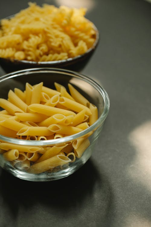Free Glass bowl of penne rigate pasta near heap of raw fusilli noodles placed on dark table in kitchen with sunlight on blurred background Stock Photo