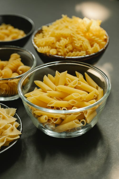 Uncooked penne rigate pasta in glass bowl near plate with fusilli placed on dark table with abundance of macaroni in kitchen