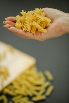 Close-up of a hand holding uncooked rotini pasta against a blurred background.