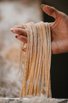 A close-up view of a hand holding freshly made pasta strings, dusted with flour.
