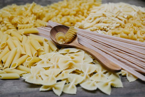 Arrangement of uncooked various pasta including spaghetti fusilli farfalle and penne heaped on table with wooden spoon