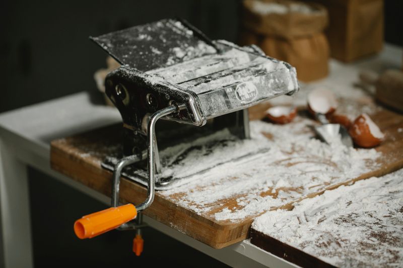 Modern manual pasta roller and cutter machine placed on wooden surface covered with flour in kitchen