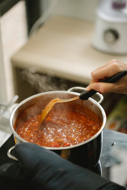 Free High angle crop faceless chef in potholder stirring bolognese sauce in pan stewing on stove in modern kitchen Stock Photo