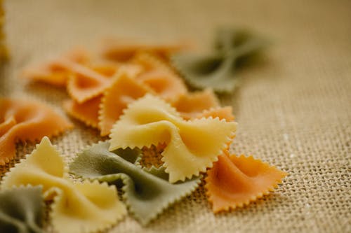 Selective focus of uncooked multicolored farfalle pasta scattered on table in light kitchen