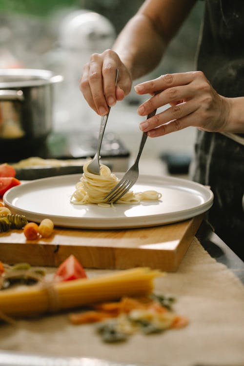 Crop anonymous female chef serving freshly cooked appetizing spaghetti on white plate while cooking in light kitchen