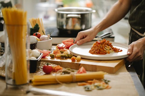 Crop anonymous cook standing at table with various ingredients and cooking pasta with meat and tomatoes