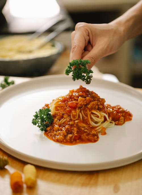 High angle of crop anonymous cook adding parsley to spaghetti with meat and sauce in kitchen