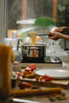 Crop anonymous housewife with tongs cooking delicious spaghetti pasta in saucepan in modern kitchen