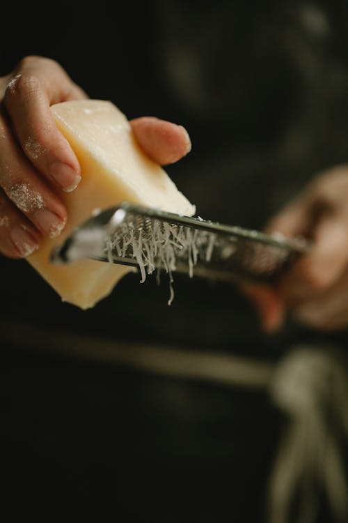 Crop unrecognizable female chef grating hard cheese on grater while cooking in kitchen