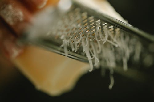 Crop unrecognizable chef grating delicious cheese on narrow stainless grater in dark kitchen