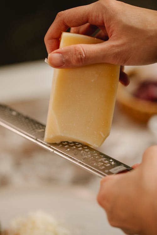 Crop anonymous chef grating hard cheese on narrow stainless grater while cooking lunch
