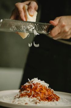 Crop anonymous female chef grating fresh cheese on yummy spaghetti with tomato sauce on white plate