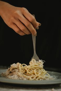 Crop anonymous female rolling yummy spaghetti in cream sauce on fork against black background