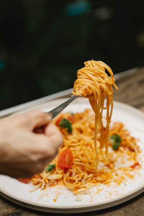 Crop anonymous person eating appetizing yummy spaghetti in tomato sauce served on white plate with parsley