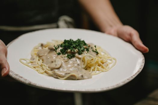 Crop anonymous female serving plate of delicious freshly cooked spaghetti with cream mushroom sauce and garnished with chopped parsley