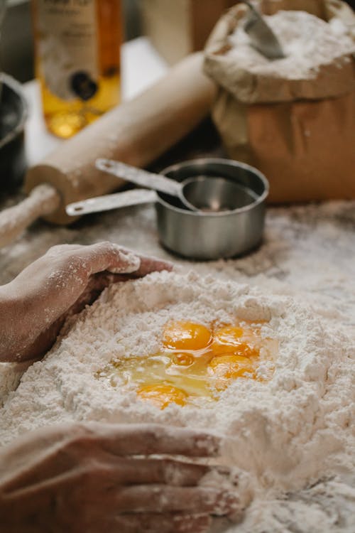 From above of crop anonymous female preparing dough with flour and eggs during cooking traditional Italian pasta standing at table with various utensils in kitchen