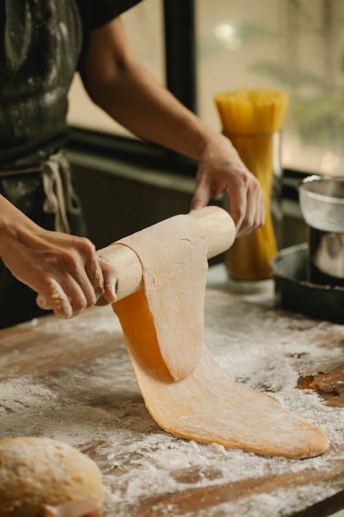 Free Crop anonymous woman in casual clothes and apron flattening homemade dough with rolling pin at floury wooden table during pasta preparation in kitchen Stock Photo