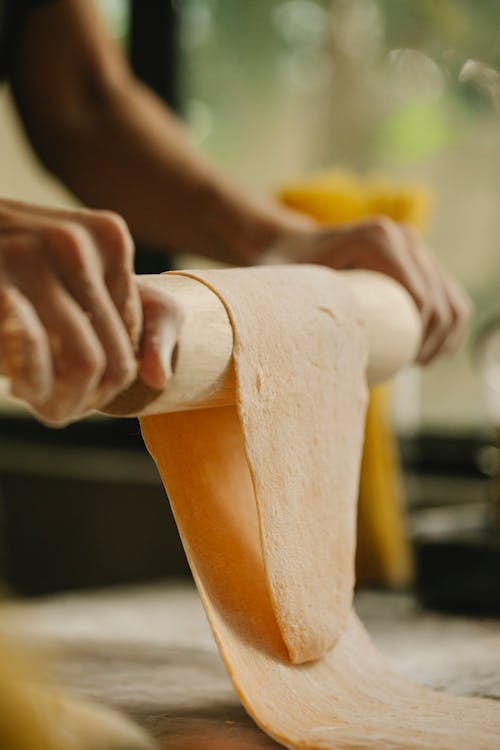 Anonymous lady flattening dough while preparing pasta in kitchen