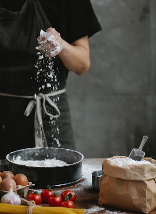 Crop anonymous female in apron adding flour into metal pan while making pasta dough standing at wooden table with eggs and tomatoes in kitchen