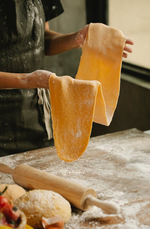 Crop anonymous female in apron showing thin dough near table with flour and rolling pin