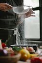 Crop anonymous female in apron sifting flour with metal sieve on table with ingredients