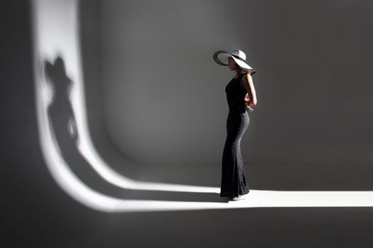 Full body of unrecognizable slim woman model in stylish long black dress and hat standing in dark studio and illuminated by light