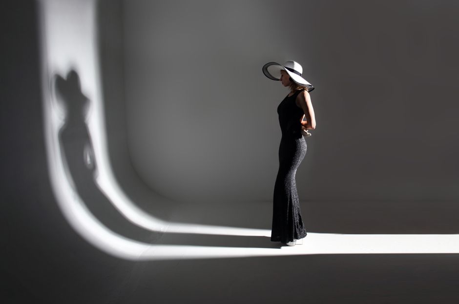 Full body of unrecognizable slim woman model in stylish long black dress and hat standing in dark studio and illuminated by light