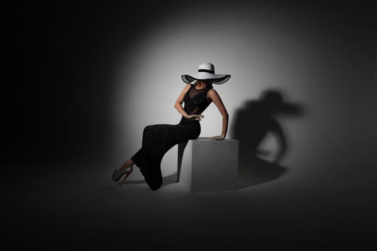 Stylish Young Lady Sitting On Cube In Studio Near Wall With Shadow