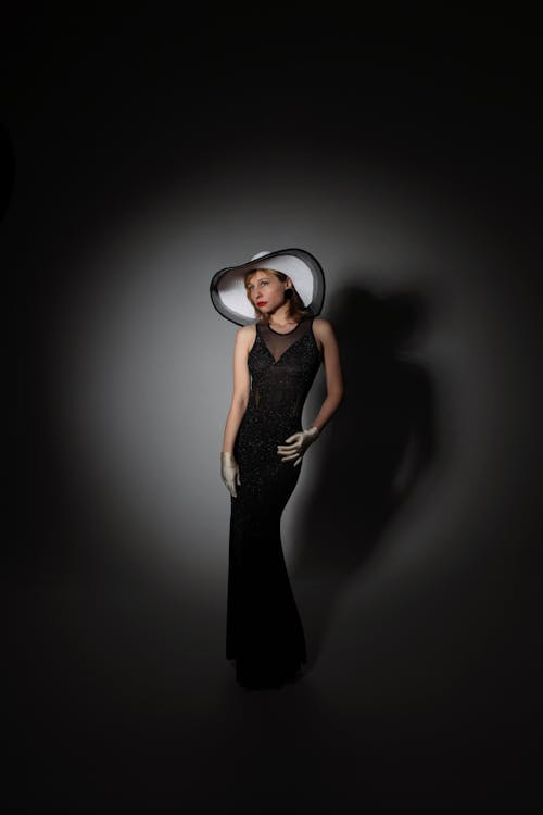 Full body of self assure young female model in elegant black dress and straw hat standing in dark studio near white wall with shadow and looking away