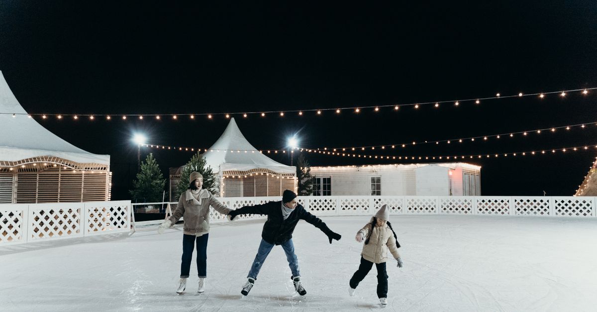 A family enjoying ice skating together on an outdoor rink at night under festive string lights.