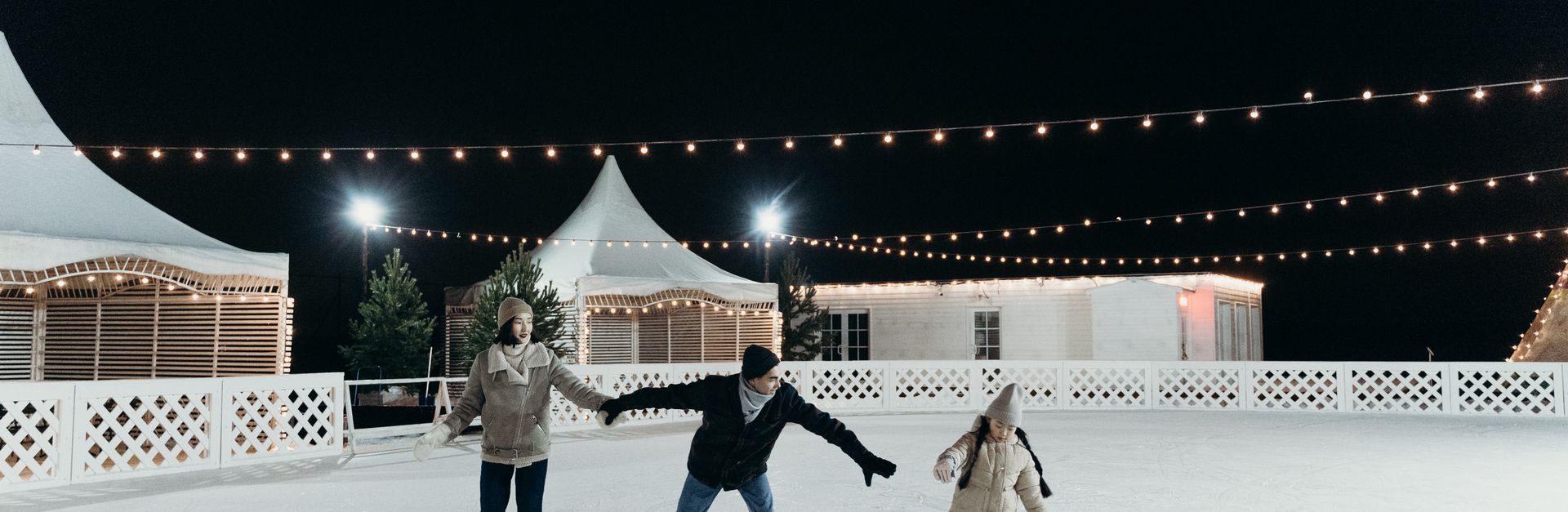 A family enjoying ice skating together on an outdoor rink at night under festive string lights.