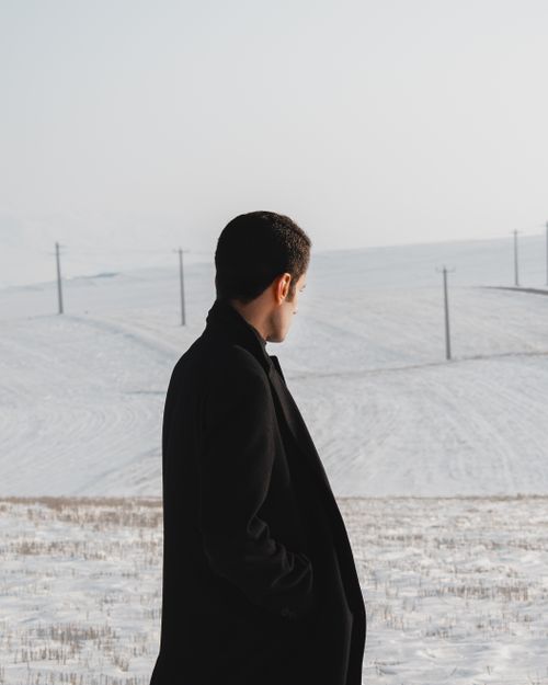Free Side view of unrecognizable young male wearing stylish warm black coat with hands in pockets standing in snowy terrain Stock Photo