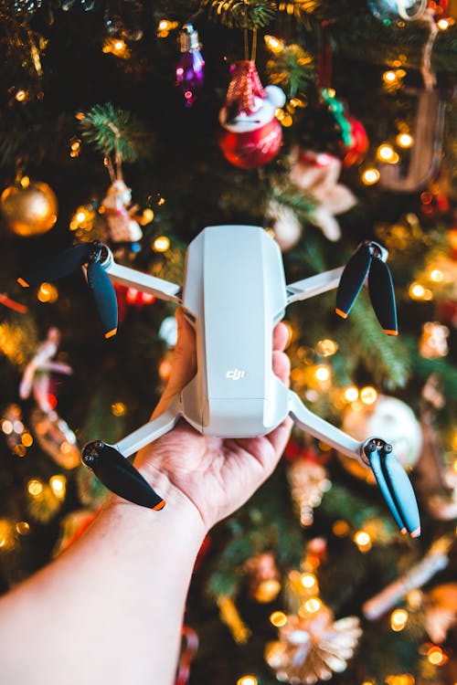 Crop unrecognizable male demonstrating modern drone near Christmas tree decorated with various baubles and glowing garlands