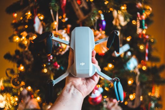 Crop faceless man holding contemporary drone in hand near Christmas tree decorated with toys and fairy lights