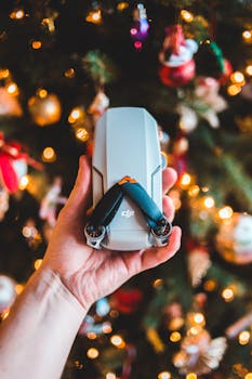 A hand holding a drone in front of a decorated Christmas tree with festive lights.