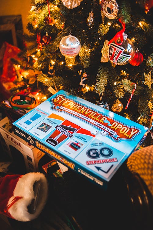 From above of boxes with various board games placed under Xmas tree with glowing fairy lights and baubles at home